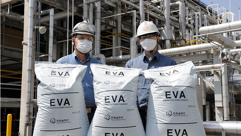 Two Hanwha Solutions employees dressed in industrial gear display bags of EVA outside of a Hanwha Solutions manufacturing plant